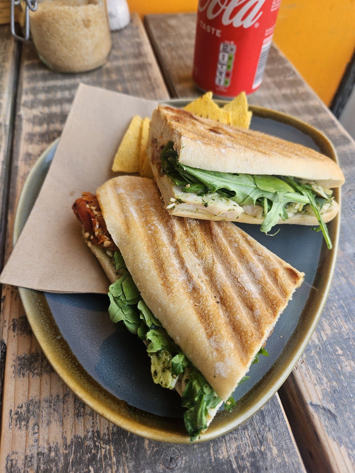 A toasted panini cut in two, filled with pesto chicken, rocket and sun-dried tomatoes, with tortilla chips, on an olive-rimmed plate on a rustic wooden table.