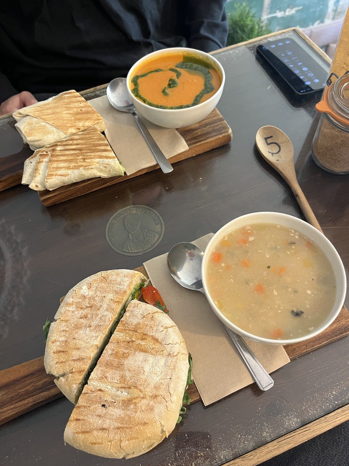 Two lunches on wooden boards: a tomato soup swirled with green herb oil beside a toasted wrap, and a chunky vegetable soup beside a toasted ciabatta, with a wooden spoon marked 5.