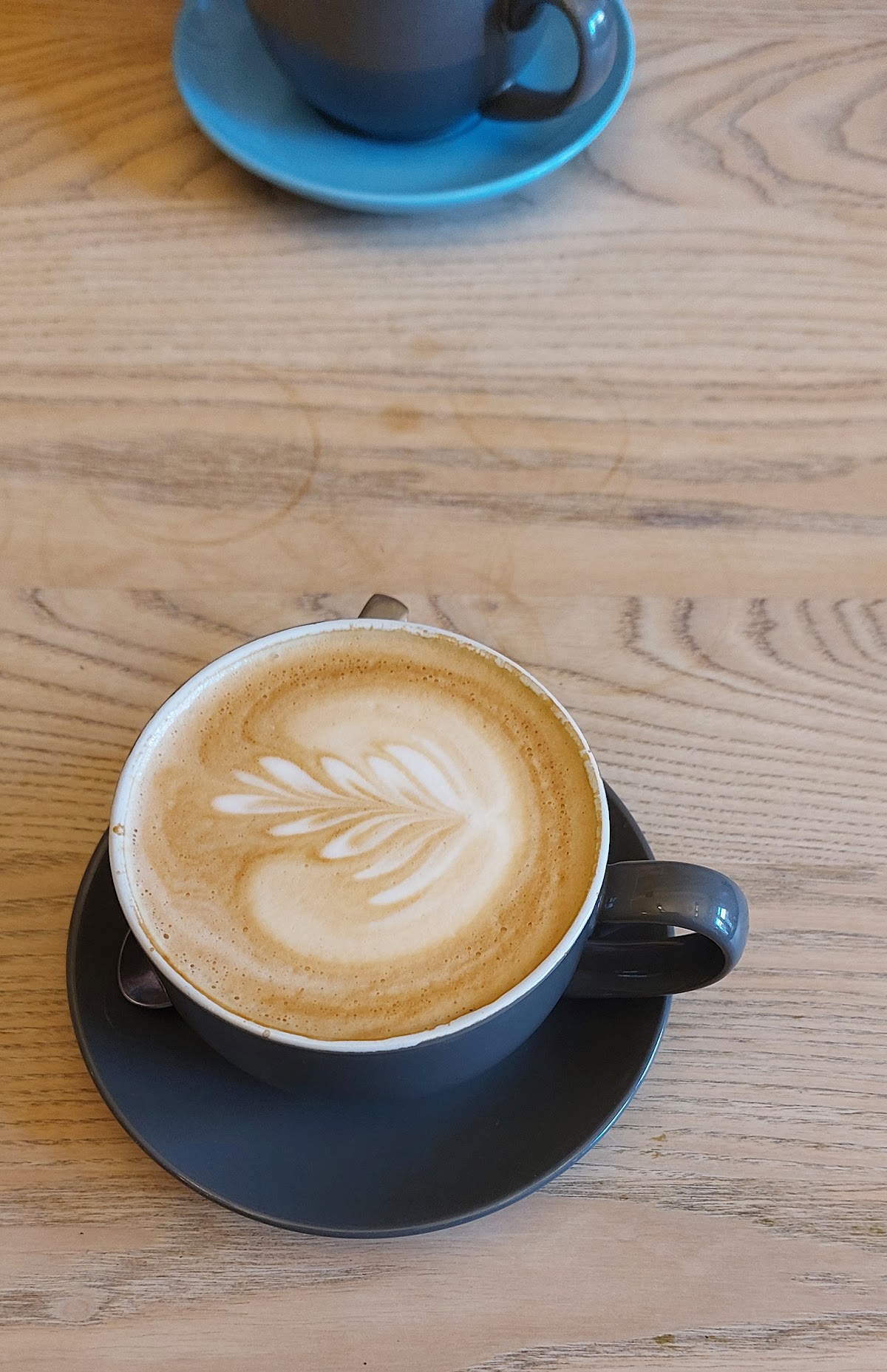 A flat white with rosetta latte art in a charcoal cup and saucer on a pale oak table, with a blue cup behind.